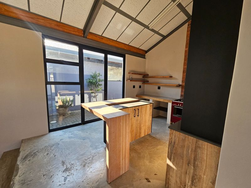 Modern industrial-style kitchen featuring a central island, exposed timber beams, concrete flooring, and large glass doors opening to an outdoor area.