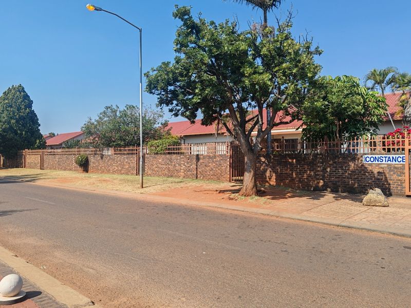 Street view of the townhouse complex featuring a brick boundary wall, metal security fencing, and mature trees along the roadside.