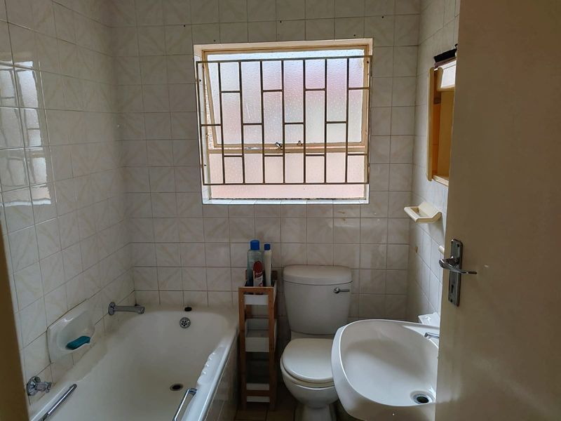 A compact bathroom featuring a bathtub, pedestal basin, and toilet, finished with dated wall tiles and fitted with security bars on the window.