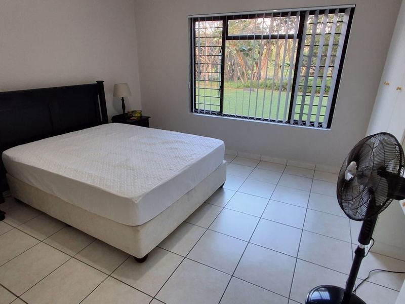 A functional bedroom featuring tiled flooring, built-in cupboards, and a large window with security bars overlooking the garden.
