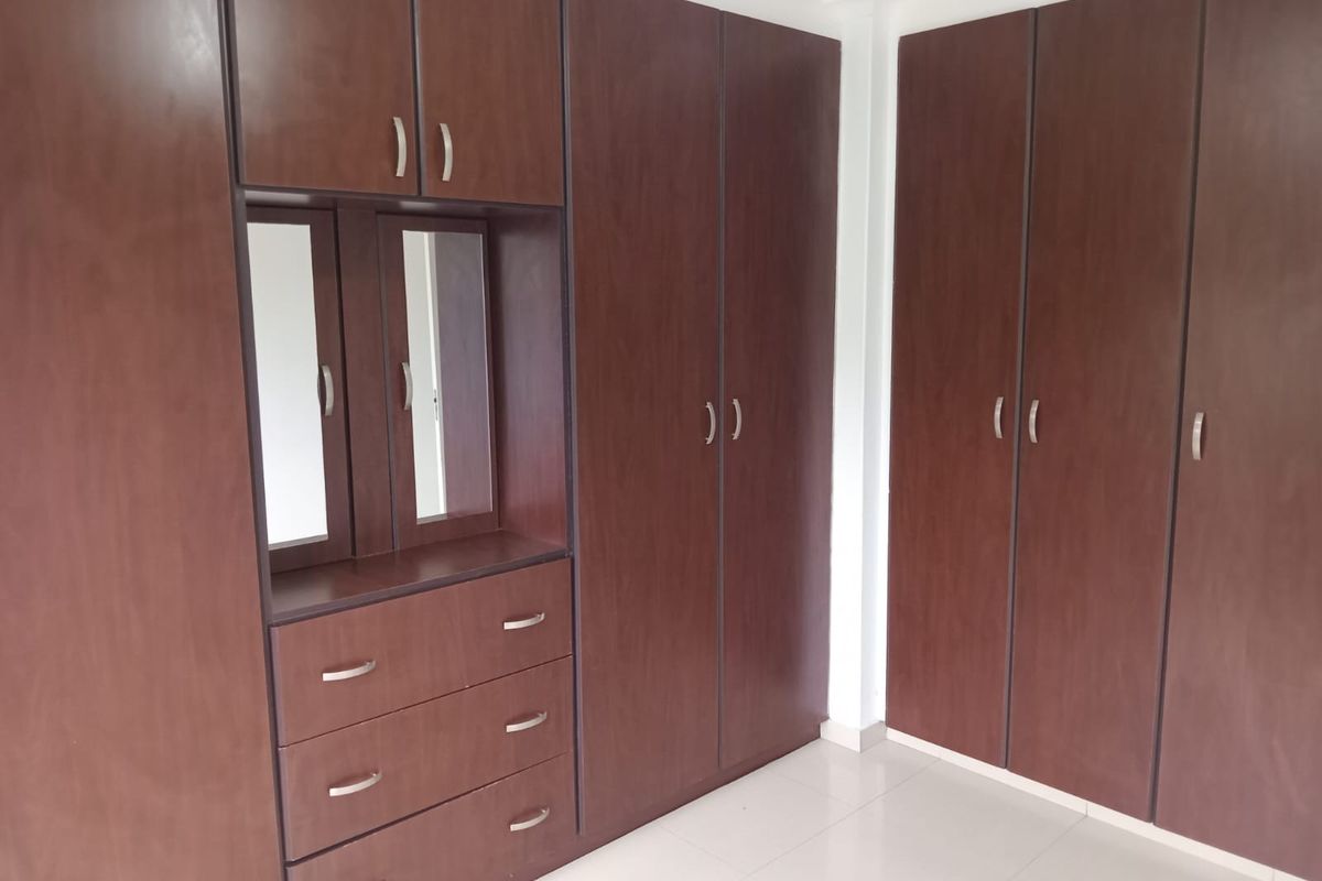 A clean bedroom featuring extensive dark wood built-in wardrobes with an integrated vanity and mirror, complemented by neutral tiled flooring.