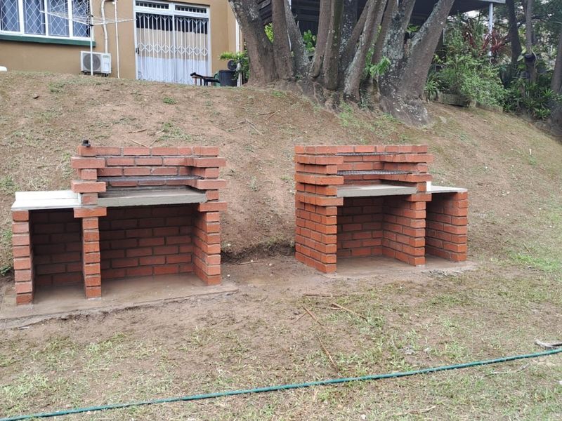 Outdoor garden area featuring two newly constructed built-in brick braai stations with concrete countertops set against a sloped lawn.