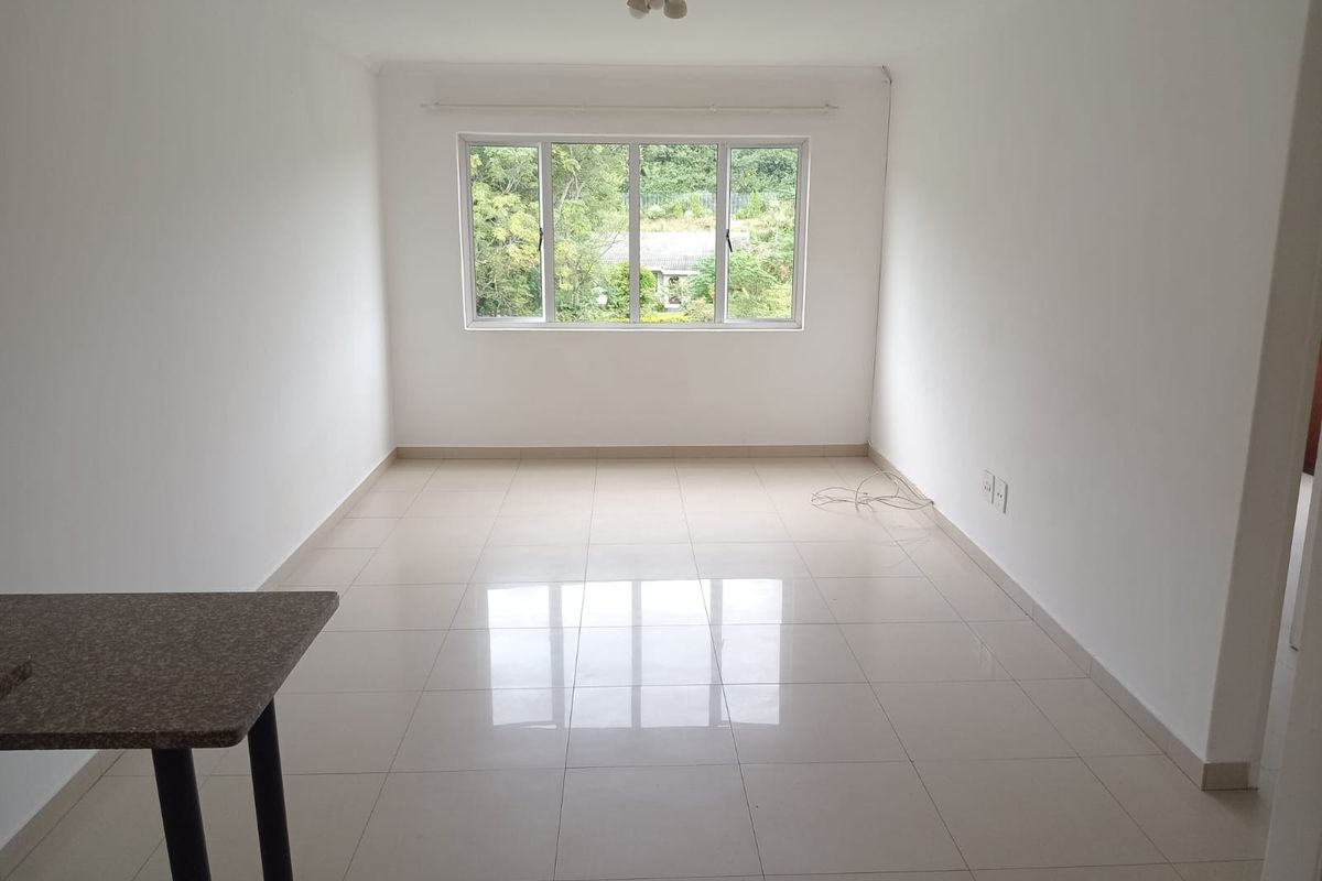 A bright, empty living room featuring neutral tiled flooring, white walls, and a large window providing natural light and a garden view.