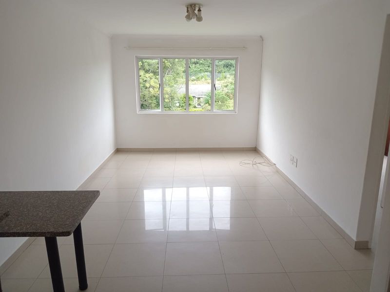A bright, empty living room featuring neutral tiled flooring, white walls, and a large window providing natural light and a garden view.