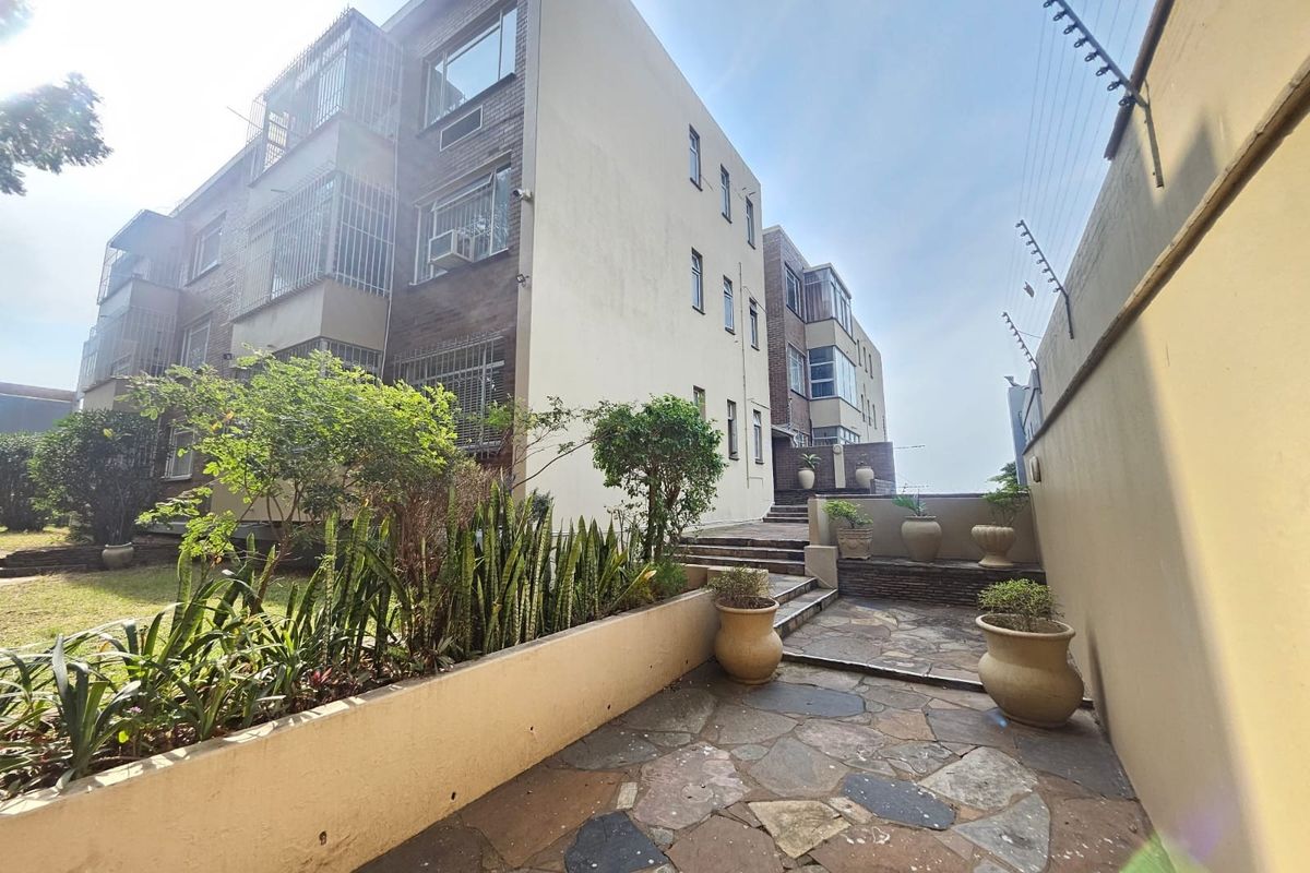 Rear exterior view of the apartment building featuring a stone-paved patio area, landscaped garden beds, and perimeter security fencing.