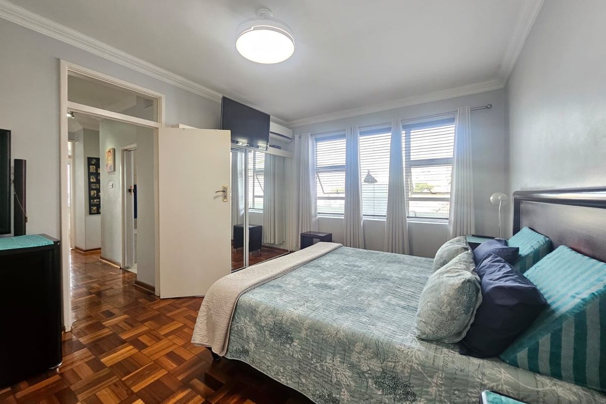 A well-maintained bedroom featuring classic parquet flooring, built-in mirrored wardrobes, an air conditioning unit, and large windows allowing for ample natural light.
