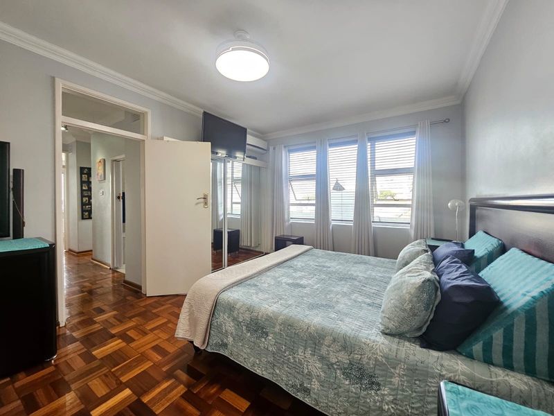 A well-maintained bedroom featuring classic parquet flooring, built-in mirrored wardrobes, an air conditioning unit, and large windows allowing for ample natural light.
