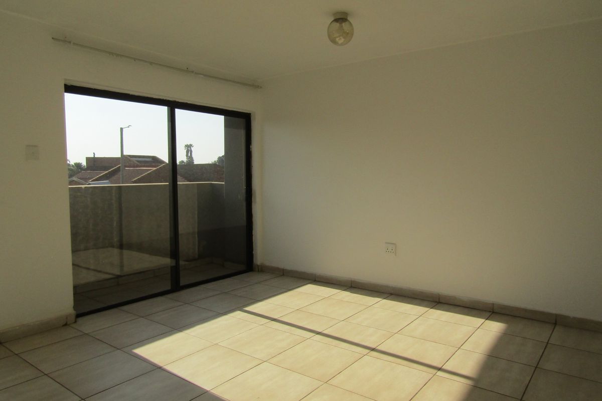 An empty, sunlit living room featuring neutral tiled flooring and a large sliding glass door providing access to an outdoor balcony area.