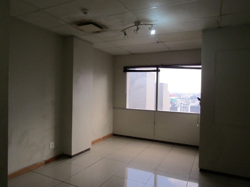 An empty bedroom featuring tiled flooring, a drop ceiling with track lighting, and a window with blinds offering an urban city view.
