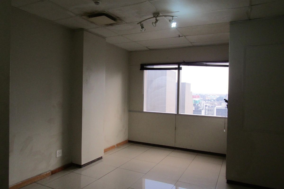 An empty bedroom featuring tiled flooring, a drop ceiling with track lighting, and a window with blinds offering an urban city view.