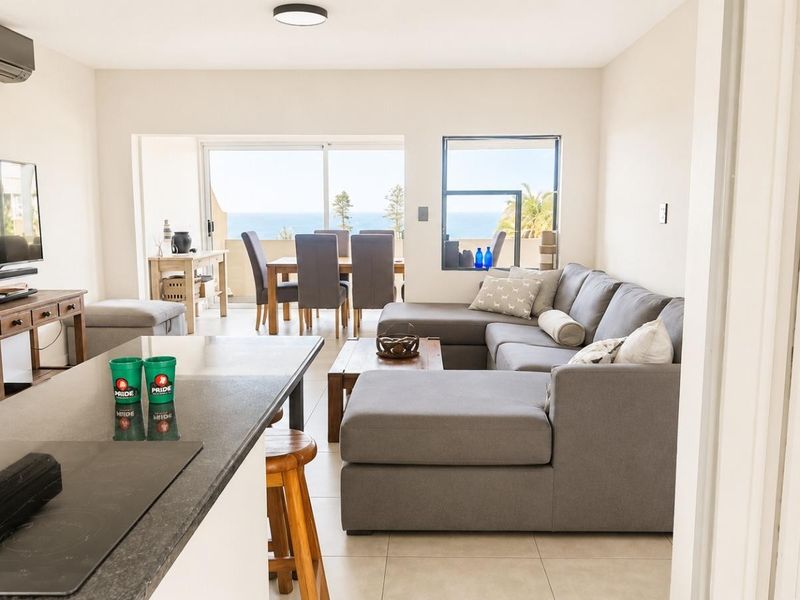 Bright open-plan living and kitchen area featuring a stone-topped island, tiled floors, and sliding doors leading to a balcony with ocean views.