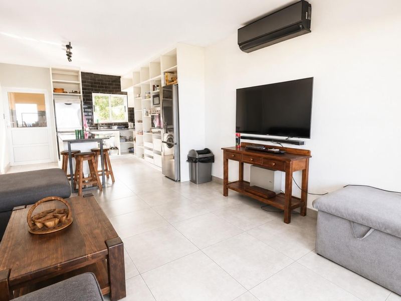 An open-plan living and kitchen area featuring tiled floors, built-in shelving, a modern kitchen with a black backsplash, and a wall-mounted air conditioning unit.