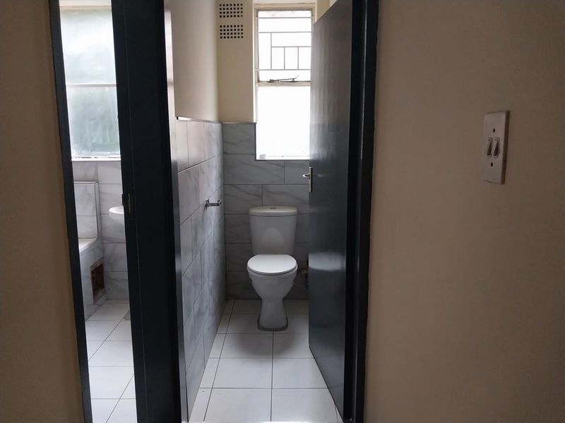 A functional bathroom featuring tiled walls and flooring, a standard toilet, and a window with security bars, showing signs of minor wear.