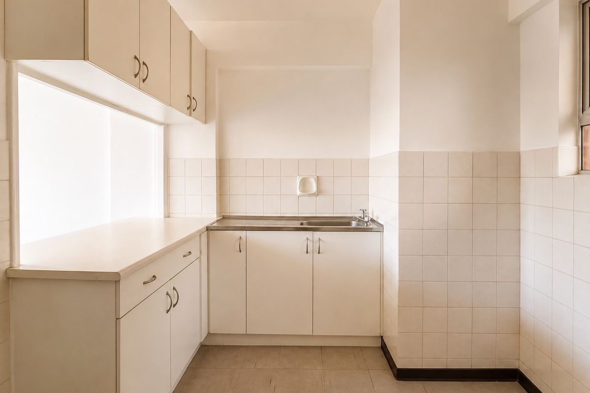 A clean, empty kitchen featuring white built-in cabinetry, a stainless steel sink, and neutral tiled splashbacks and flooring.