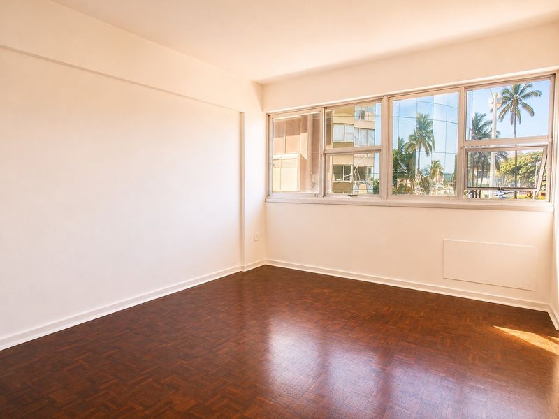 A bright, empty bedroom featuring classic parquet flooring and large windows that provide ample natural light and an urban view.