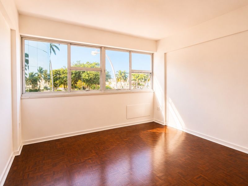 A bright, empty bedroom featuring polished parquet flooring and a large window providing natural light and exterior views.