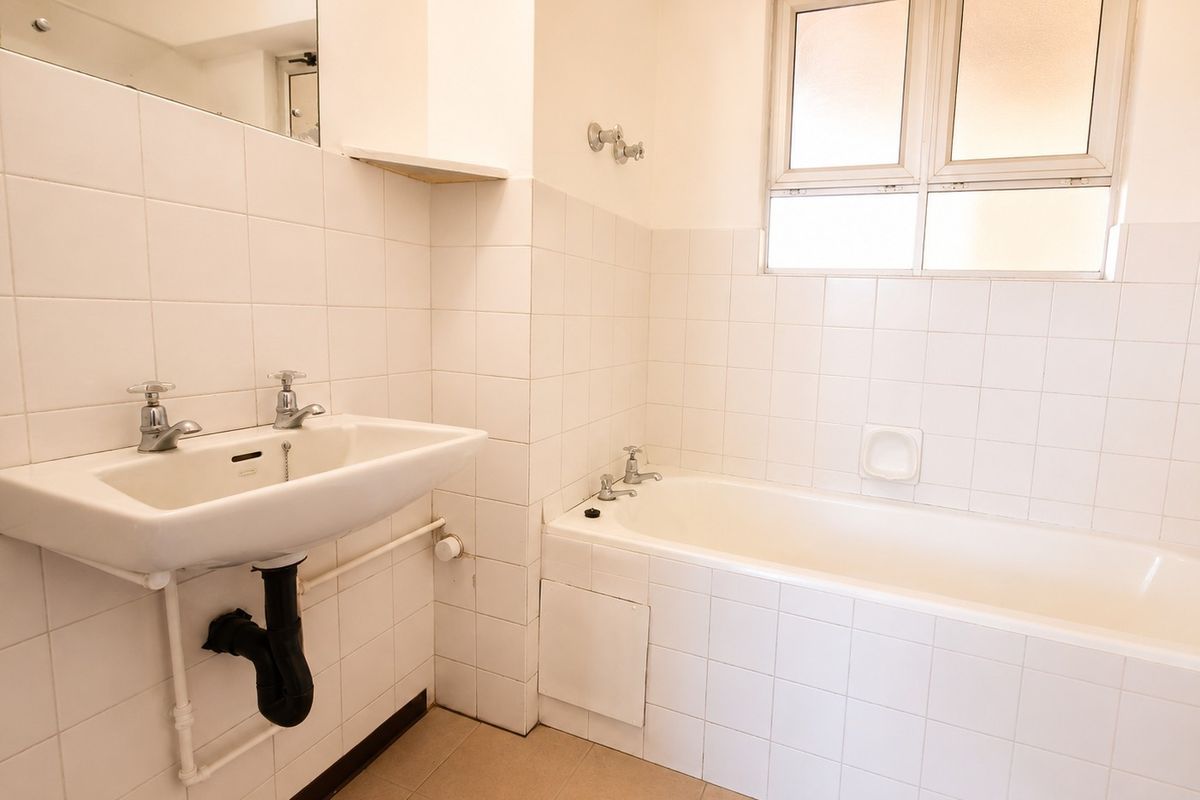 A functional, neutral-toned bathroom featuring a built-in bathtub, wall-mounted basin, and tiled finishes, offering a clean canvas for modern updates.
