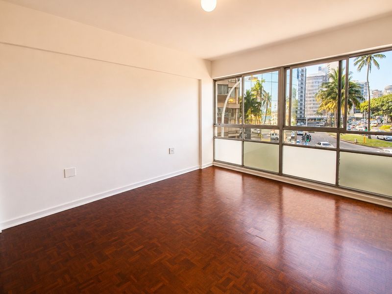 A bright, empty living area featuring polished parquet flooring and large windows offering an urban street view.