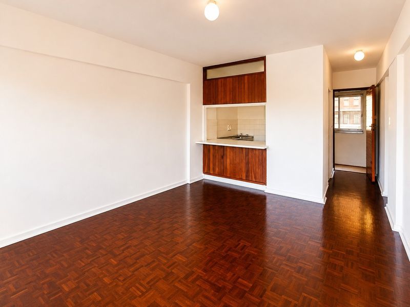 An open-plan living area featuring polished parquet flooring, neutral walls, and a built-in wooden cabinetry unit with a kitchen pass-through window.