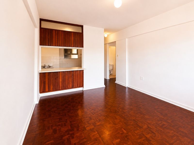 An open-plan living area featuring original parquet flooring and a timber-paneled kitchen servery, leading to a hallway and bathroom.
