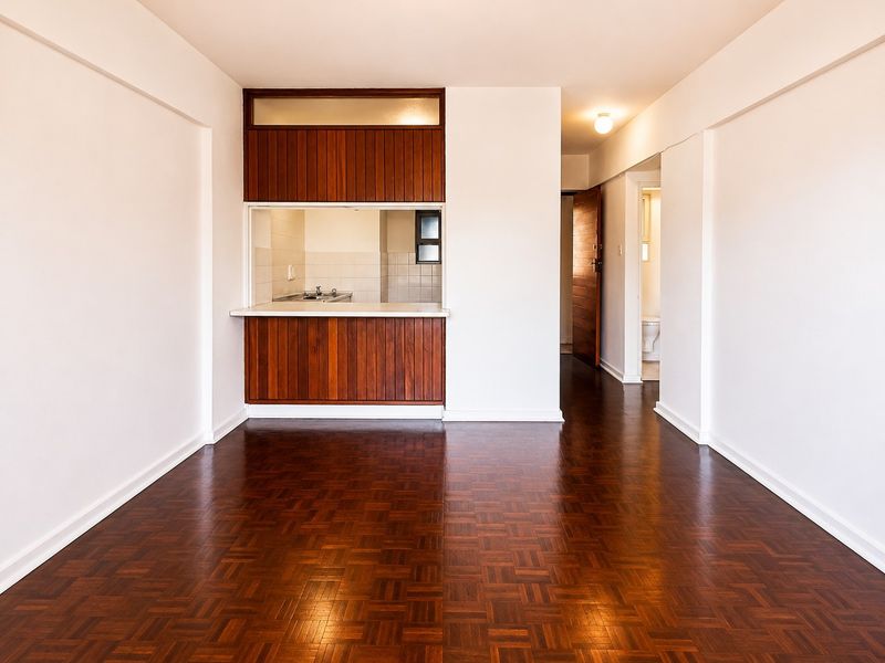 An open-plan living area featuring classic parquet flooring and a timber kitchen servery, leading to a hallway and bathroom.