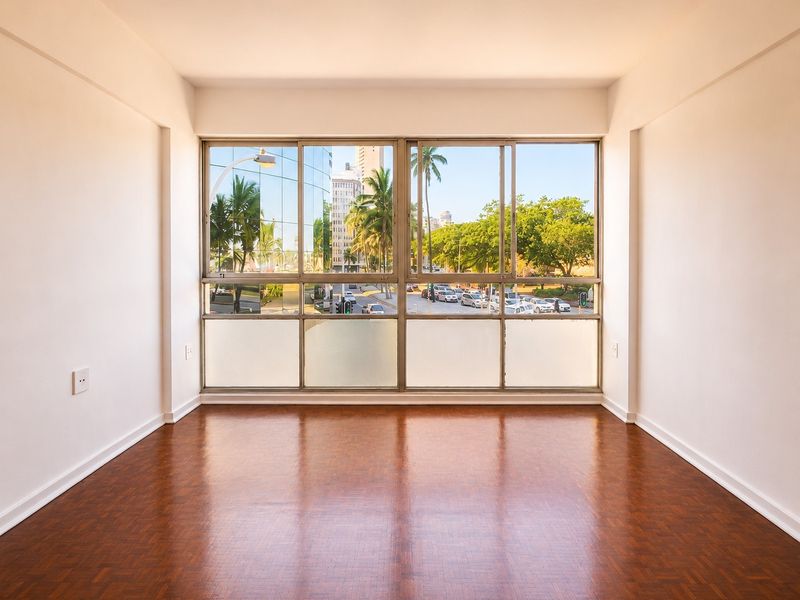 A bright, empty living room featuring polished parquet flooring and large sliding windows with an urban street view.