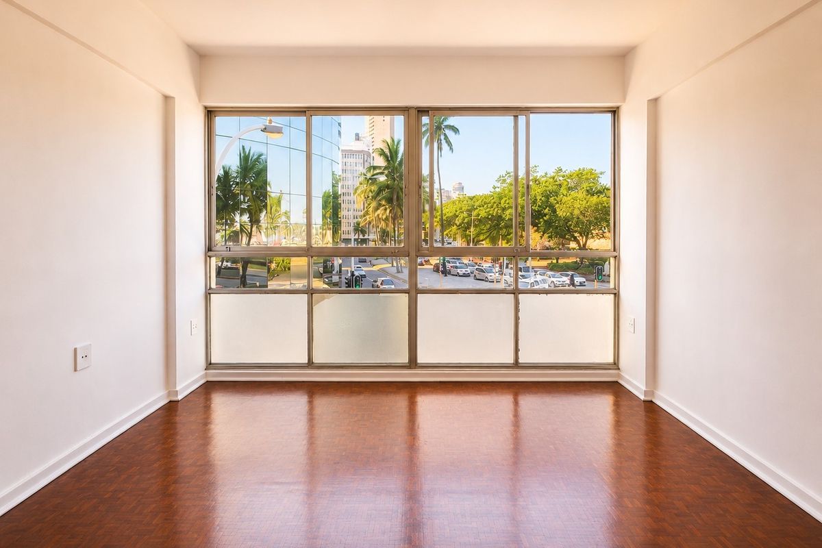 A bright, empty living room featuring polished parquet flooring and large sliding windows with an urban street view.