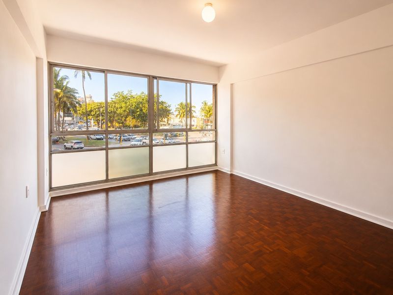 A bright, empty living area featuring polished parquet flooring and large windows that provide ample natural light and an urban view.