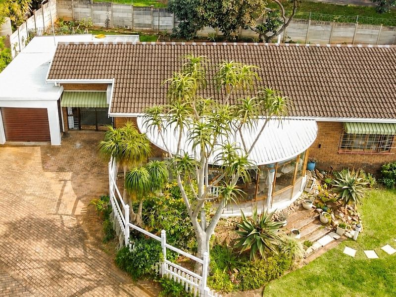 Aerial view of a townhouse featuring a tiled roof, brick exterior, paved driveway, single garage, and a landscaped garden with a curved patio area.