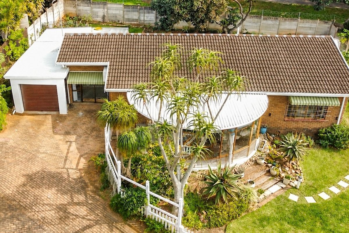 Aerial view of a townhouse featuring a tiled roof, brick exterior, paved driveway, single garage, and a landscaped garden with a curved patio area.