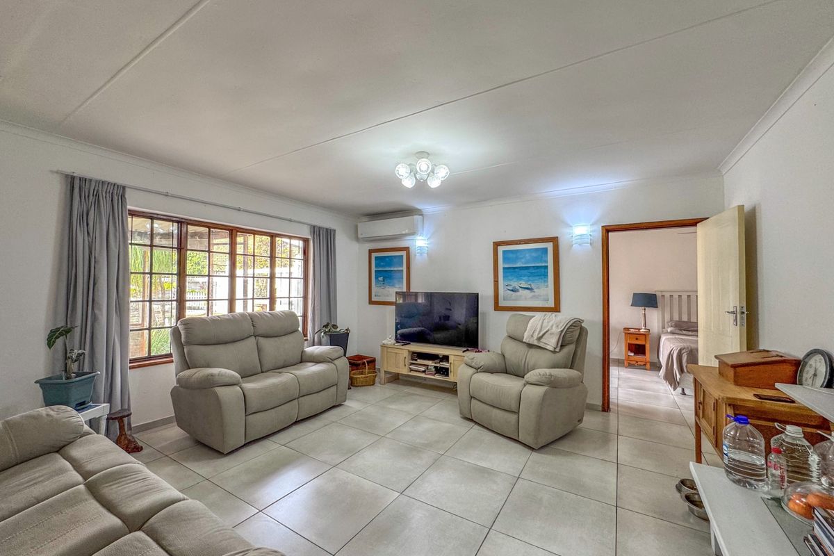 A spacious living room featuring neutral tiled flooring, a wall-mounted air conditioning unit, and large timber-framed windows allowing for natural light.