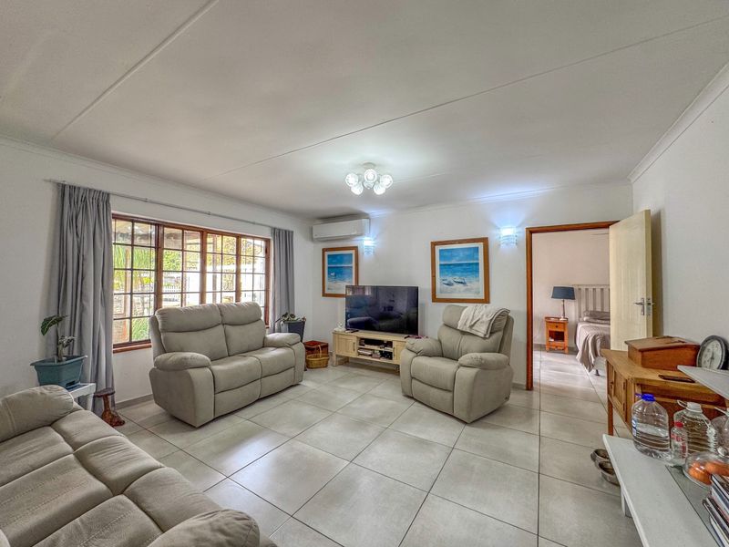 A spacious living room featuring neutral tiled flooring, a wall-mounted air conditioning unit, and large timber-framed windows allowing for natural light.