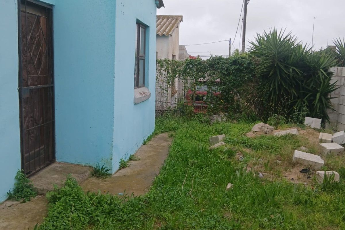 Rear exterior view of the property featuring a concrete pathway, security gate, and an overgrown garden area requiring maintenance and landscaping.