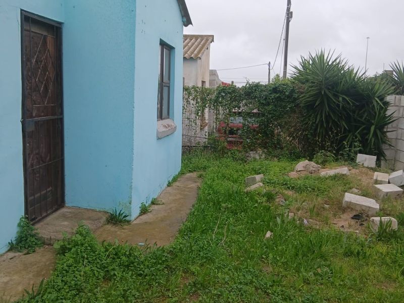 Rear exterior view of the property featuring a concrete pathway, security gate, and an overgrown garden area requiring maintenance and landscaping.