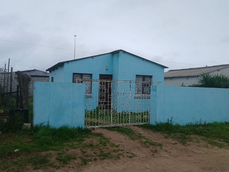 Front view of a single-story detached house featuring a perimeter wall, wrought iron gate, and metal roof, set on an unpaved access road.