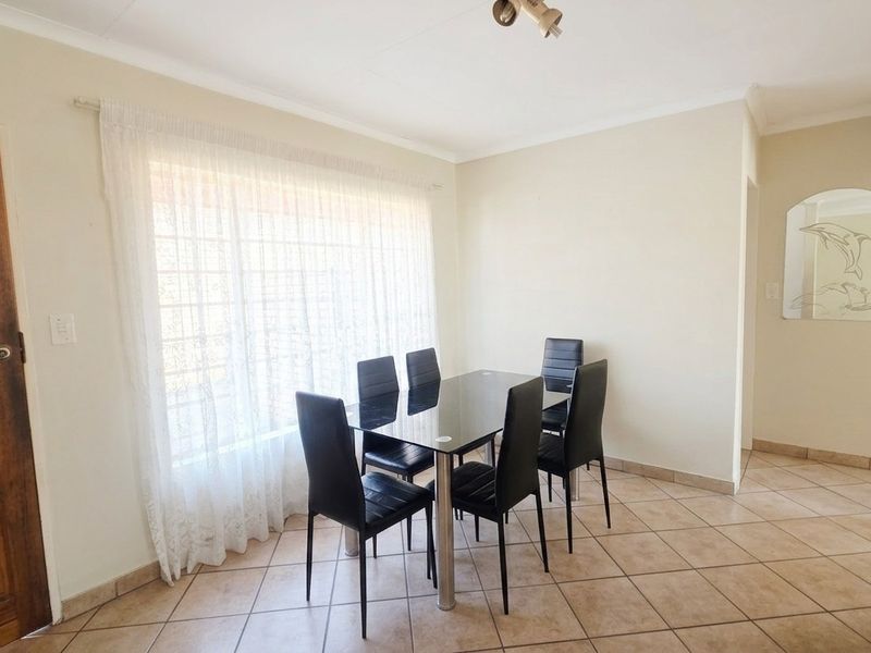 A dining area featuring tiled flooring, a large window with security bars, and a wooden door, providing a functional space for meals.