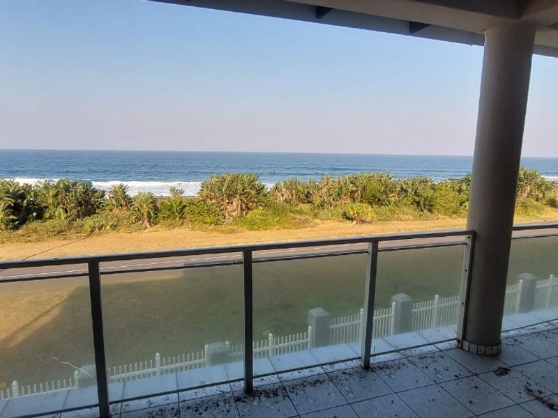 A spacious covered balcony featuring a glass balustrade and concrete columns, offering an expansive, unobstructed view of the ocean and coastal landscape.