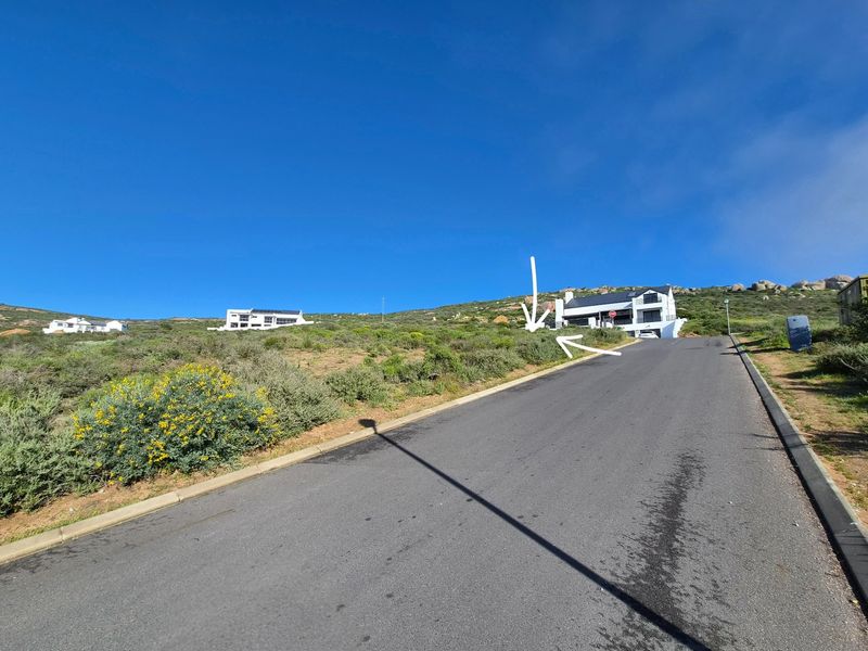A street-level view of a sloped residential road leading towards a modern property on a hillside, showcasing the surrounding landscape and neighborhood approach.