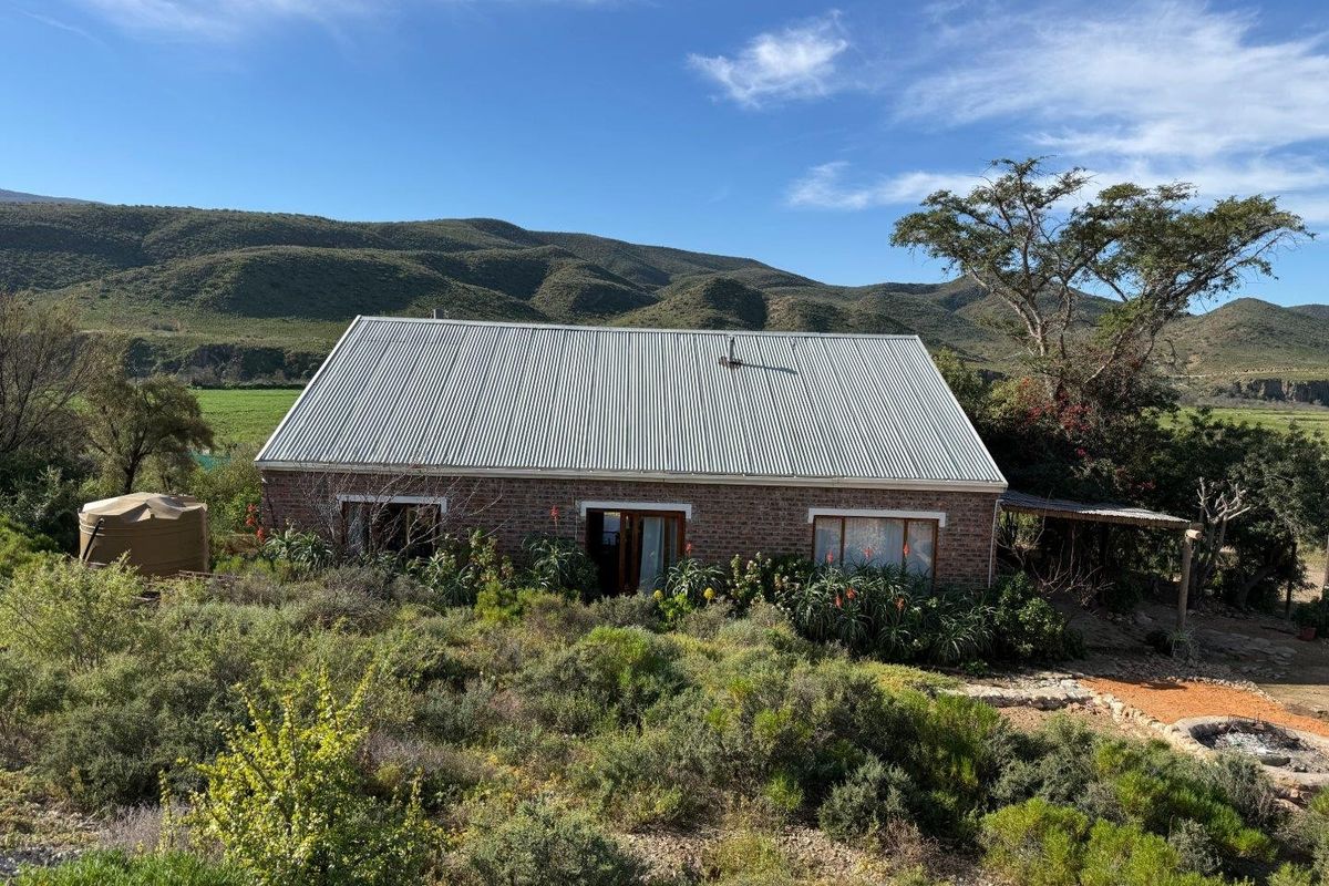 A rustic face brick farmhouse featuring a corrugated metal roof, set against a scenic mountain landscape with natural vegetation and a water storage tank.