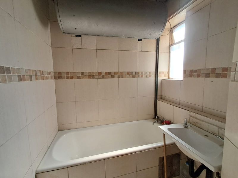 A dated bathroom featuring a standard bathtub, wall-mounted basin, and exposed water heater unit with full-height wall tiling.