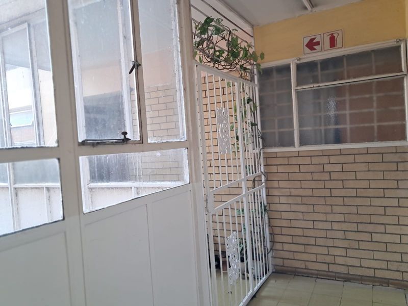 A functional apartment hallway featuring a security gate, brick wall finishes, and steel-framed windows, providing access to the unit entrance.