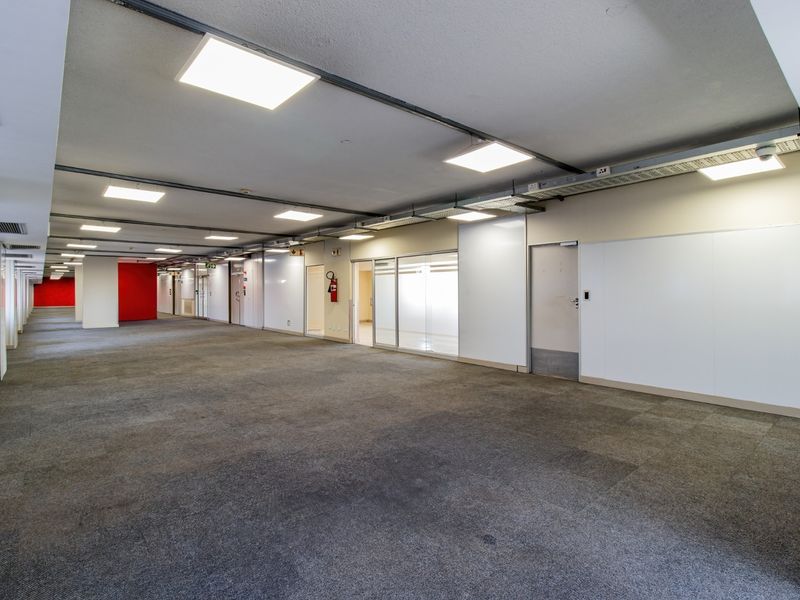 A spacious, open-plan commercial office area featuring carpeted flooring, recessed LED lighting, exposed cable management systems, and glass-partitioned office suites.