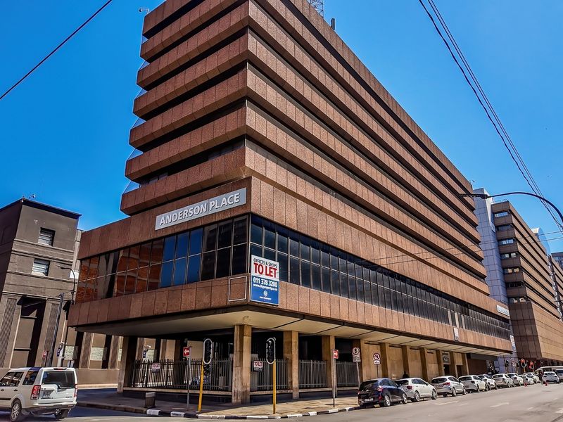 A multi-story commercial office building featuring a prominent brown facade, large glass windows, and ground-level security fencing located on a street corner.