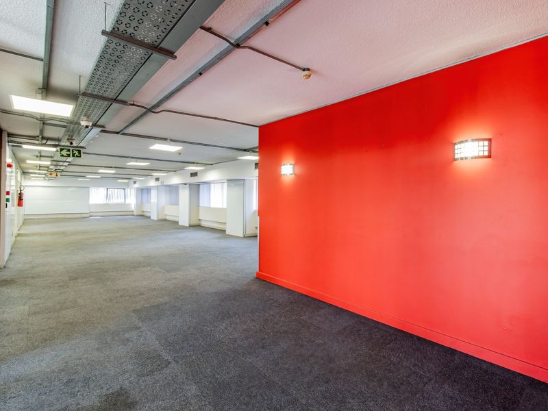 An open-plan commercial office space featuring exposed industrial cable trays, recessed lighting, and a prominent red feature wall.