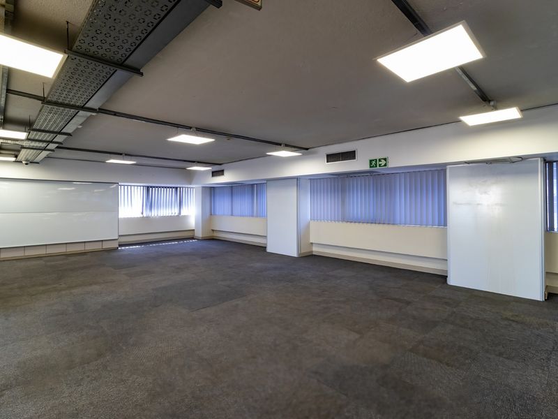 Spacious open-plan commercial office area featuring carpeted flooring, exposed cable management, and large windows with vertical blinds.