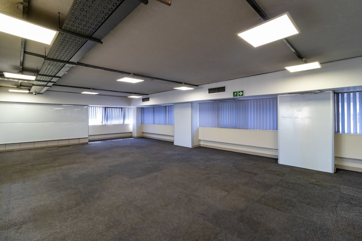 Spacious open-plan commercial office area featuring carpeted flooring, exposed cable management, and large windows with vertical blinds.