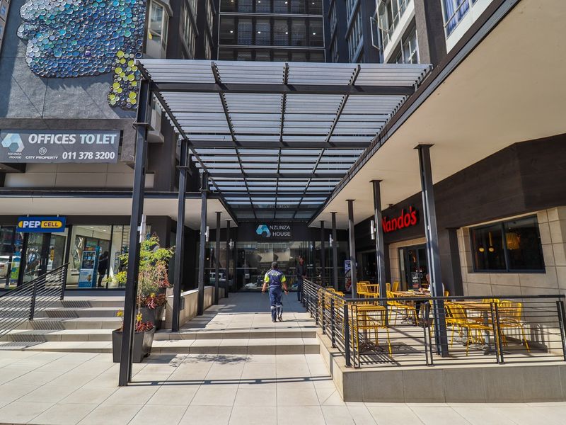 Modern commercial building entrance featuring a covered walkway, paved access, and integrated retail storefronts with metal structural elements.