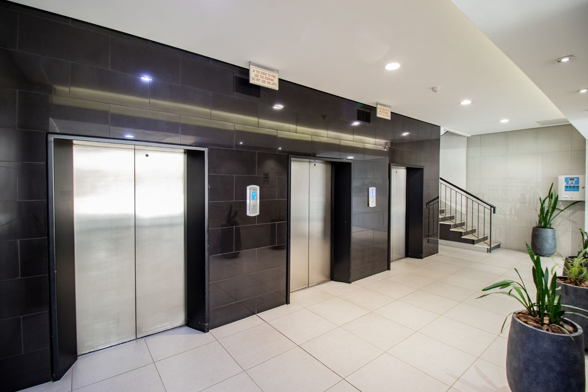 A modern commercial building lobby featuring a bank of stainless steel elevators, tiled flooring, and a staircase with a metal railing.