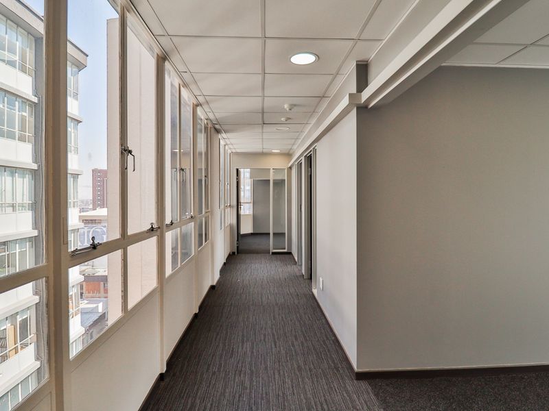 A bright, modern commercial hallway featuring carpeted flooring, a suspended ceiling with recessed lighting, and large windows providing ample natural light.