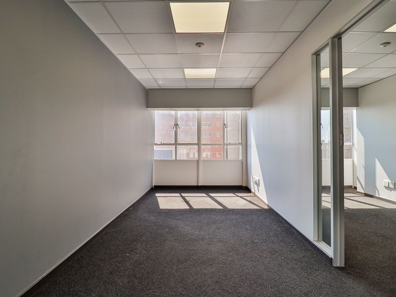 A bright, empty commercial office space featuring a suspended ceiling, recessed lighting, carpeted flooring, and a glass partition wall.